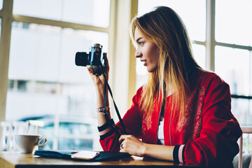 Professional female photographer taking picture on old camera focusing lens concentrated on work.Hipster girl fond of photography using vintage equipment making images out of window sitting in cafe