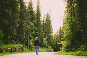 Fototapeta premium The concept of walking alone. Young caucasian blond man in jeans walks along a road in a forest park. A cozy image in calm warm colors.