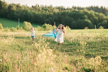 Fototapeta premium Cute curly little girl in white dress and boy in white shirt, running on sunset meadow with kite. Healthy, enjoy countryside summer and happy childhood concept