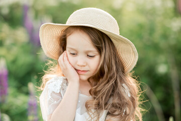 Portrait of cute curly little girl in white dress and hat with brim, staying in the garden. Healthy, enjoy countryside summer and happy childhood concept