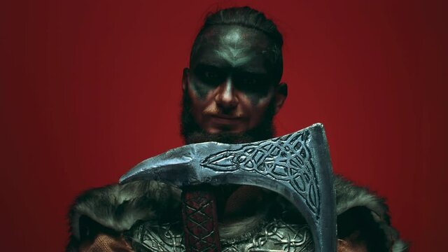 A Viking in war paint leans his beard on an axe, opens his eyes and looks aggressively at the camera. Menacing Viking man on a red background