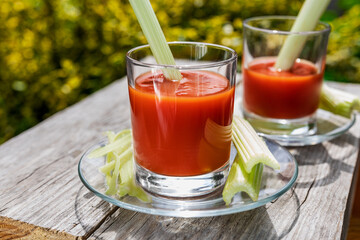 Two glasses of tomato juice with parsley and celery decorations, stand in the open air on wooden boards, the rays of the sun shine, shallow depth of field, selective focus. The concept of natural