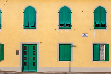 Fototapeta premium Manzano, Italy - 25.04.2020: old yellow house facade with green wooden door and green wooden window shutters. The doll looks out the one window. Board on building: 