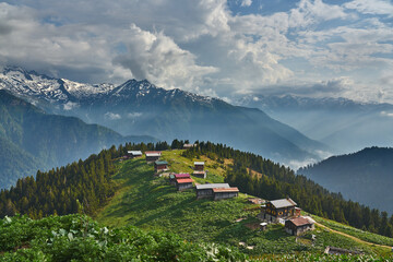 Landscape photo of Pokut Plateau with traditional wooden houses, snowy mountains, clouds and forest. Taken in summer at northeastern Black Sea / Karadeniz region of Turkey