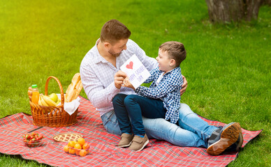 I love you daddy. Adorable boy giving greeting card to father during family picnic on green meadow