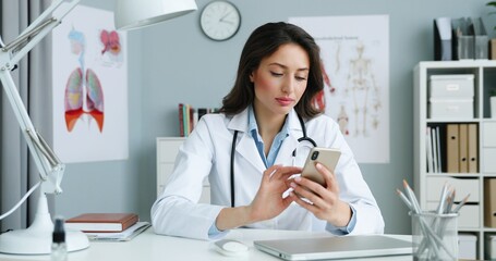 Caucasian young pretty female doctor in white gown sitting at desk in cabinet and using smartphone and texting message. Woman medic scrolling, tapping sms and browsing on mobile phone online.