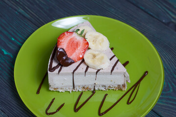 Cheesecake on a plate. Garnished with strawberries, bananas and chocolate. On brushed pine boards painted black and green.