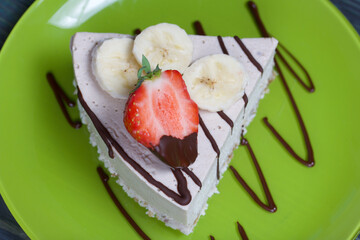 Cheesecake on a plate. Garnished with strawberries, bananas and chocolate. On brushed pine boards painted black and green.