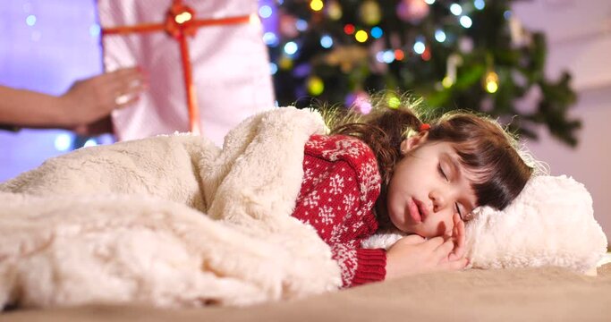   Little European brunette  girl in a burgundy sweater with a pattern sleeping  behind a white fur blanket near the Christmas tree. In the background, mom&rsquo;s hands are laying a box with a gift. 