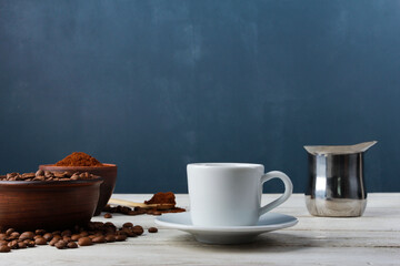 Obraz na płótnie Canvas White espresso cup, Arabica beans in clay bowls, ground powder, metal Turkish pot on while table against dark blue wall. Side view, copy space. Coffee shop, morning, baristas workplace concept