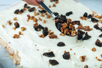 Woman spreads prune and nut filler on meringue cake on a white vintage wooden table.
