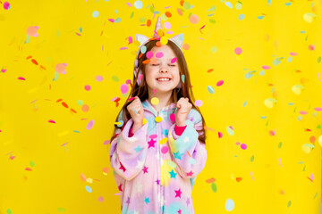 Obraz na płótnie Canvas little girl with long hair wearing a kigurumi and headphones in the shape of a unicorn is happy catching confetti standing on a yellow background