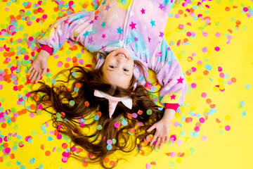 Fototapeta premium little girl with long hair wearing a kigurumi and headphones in the shape of a unicorn is happy catching confetti standing on a yellow background