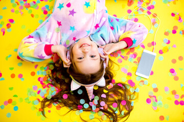 Fototapeta premium happy little girl in a unicorn kigurumi lies on a yellow background amid multicolored confetti listening to music