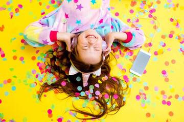 Fototapeta premium happy little girl in a unicorn kigurumi lies on a yellow background amid multicolored confetti listening to music