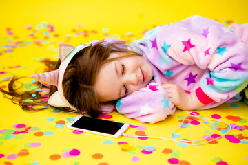 Obraz na płótnie Canvas happy little girl in a unicorn kigurumi lies on a yellow background amid multicolored confetti listening to music