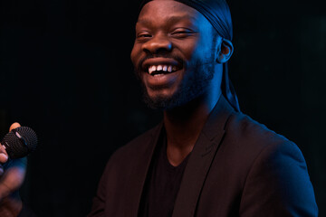 Fototapeta premium dark-skinned handsome guy in a bandana, black classic jacket and t-shirt holds a microphone in his hand, smiles and looks in the camera