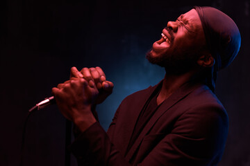 dark-skinned handsome guy in a bandana, black classic jacket and t-shirt holds a microphone in his hands and emotionally sings in a dark studio with red light