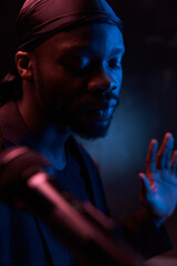 dark-skinned handsome guy near microphone in a bandana, black classic jacket and t-shirt looks down in a dark studio with blue light