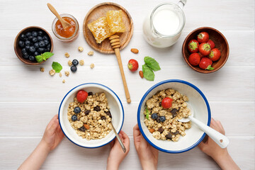 Kid's hands holding bowls with granola. Berries, milk and honeycombs on  white wooden background
