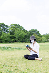 Naklejka na ściany i meble 芝生で帽子をかぶりマスクをして読書する1人の女性