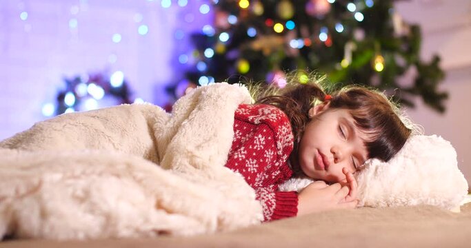   Little European brunette  girl in burgundy sweater with pattern sleeping  behind white fur blanket near the Christmas tree. In background, Santa&rsquo;s hands in red gloves are laying box with gift. 