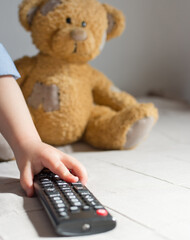 Baby hand holding the TV remote control, teddy bear in the background, selective focus