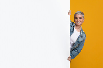 Beautiful middle-aged woman peeking from behind white advertisement board with free space