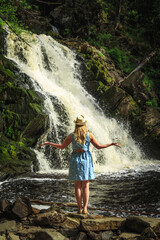 Woman in hat meditates near forest waterfall.