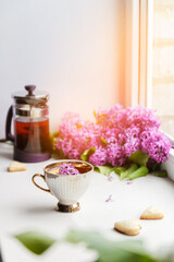 Obraz na płótnie Canvas Fresh hot black tea in a beautiful porcelain cup with a lilac bouquet in the background on a white windowsill