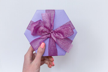 A beautiful lilac hexagon gift box with ribbon and bow on the woman hand on the white background. Flat lay. Place for text. Present