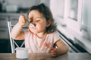 Cute, funny baby girl eating yogurt in the kitchen in a pink dress in the afternoon
