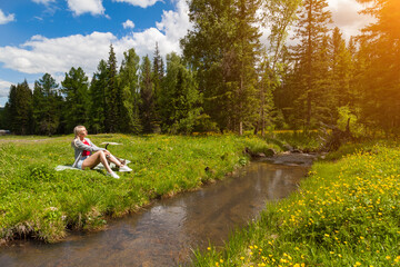 A blonde girl sits in a clearing against a forest and blue sky next to a stream, her eyes closing in the sun, a cat rubs against her legs. Halt during a walk in the Altai mountains.