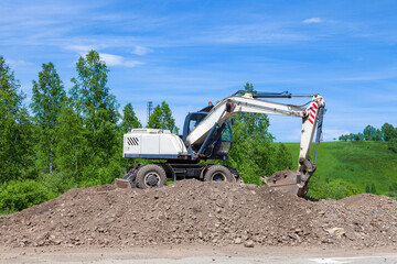 Fototapeta premium A working white escalator with a blue cabin with a lowered bucket raking on a large pile of land against a background of green trees and sky during the construction of a suburban highway.