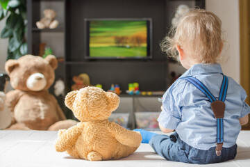 Cute baby boy and his teddy bear watching TV sitting on a couch in the living room at home