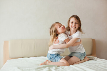 Sisters girls having fun hugging laughing on the big bed in the bedroom in a real interior.