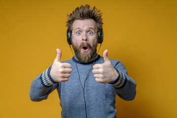 Pleasantly surprised man in headphones listens to music and makes an approving gesture, thumbs up. Yellow background. - obrazy, fototapety, plakaty