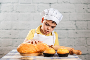un ni&ntilde;o vestido con gorro y delantal de panadero toca el pan con curiosidaden la sesion de fotos con el pan y las magdalenas. escena de aire rustico. estilo de vida, panader&iacute;a.
