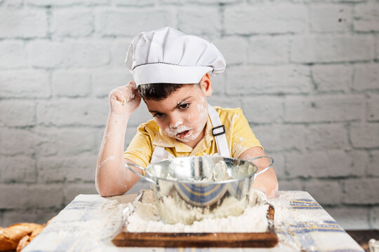 un niño vestido con gorro y delantal de panadero agarra harina con la mano, tiene la boca y cara sucia. escena de aire rustico. estilo de vida, panadería.  amasar harina para hacer pan.