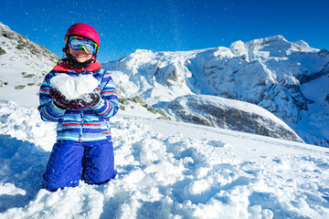 Fototapeta na wymiar Smiling happy cute girl in ski outfit mask and helmet show love concept holding snow shape in heart standing on knees with mountains on back