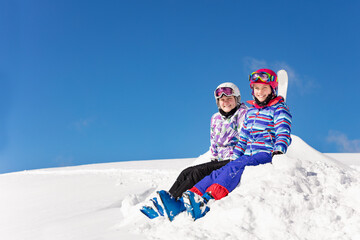 Two beautiful girls sit on pile of snow over blue background in sky outfit color mask and glasses look at camera smiling
