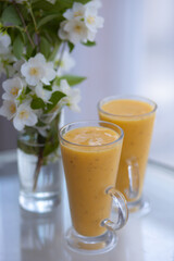two yellow smoothie cocktails in high glasses on a glass breakfast table in the morning with white flowers. Side view. Summer Spring