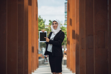 Pointing on tablet screen. Beautiful muslim successful businesswoman portrait, confident happy CEO, leader, boss or manager. Using devices, gadgets, working on the go, looks busy. Inclusive, diversity