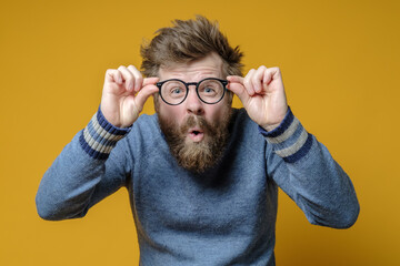 Strange shaggy, bearded man in an old sweater holds glasses in hands and squints, he saw something interesting and was surprised.