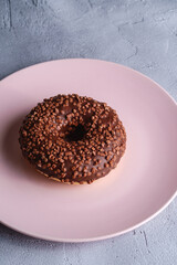 Chocolate donut with sprinkles on pink plate, sweet glazed dessert food on concrete textured background, angle view