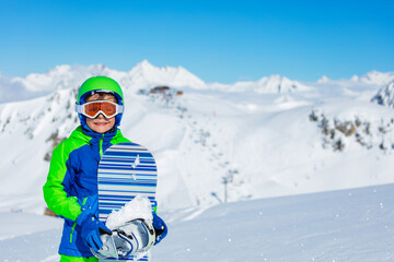 Fototapeta na wymiar Close portrait of a boy in sport helmet hold snowboard glasses over bright sunny mountain snow tops on background