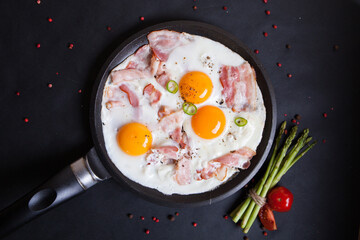 Fried eggs with three bright yolks, bacon and herbs in a black pan on a black background