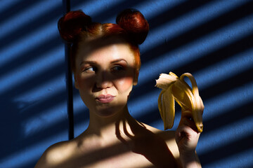 Creative portrait of a red-haired girl with a banana in her hands on a blue background with a stripe from the sun's shadow - 355212735