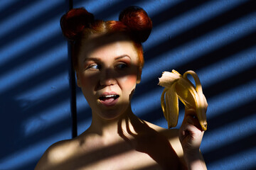 Creative portrait of a red-haired girl with a banana in her hands on a blue background with a stripe from the sun's shadow - 355212745
