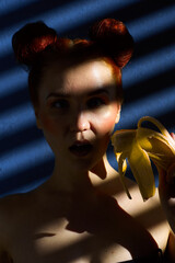 Creative portrait of a red-haired girl with a banana in her hands on a blue background with a stripe from the sun's shadow - 355212778